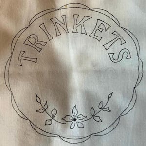May include: A white fabric circle embroidered with the word "TRINKETS" in black capital letters. The circle has a scalloped edge and a floral and leaf design at the bottom.