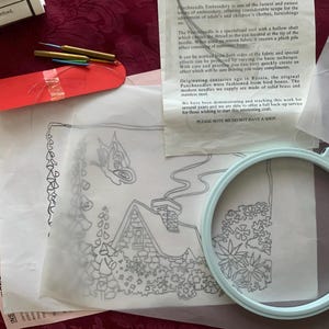 May include: A punch needle embroidery kit featuring a light blue plastic hoop, a printed pattern on translucent paper showing a house and butterfly, three brass needle tools, and an instructional sheet with text about punch needle embroidery.