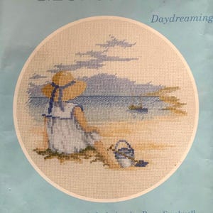 May include: Cross-stitch embroidery depicting a person on a beach, wearing a hat and dress, with a bucket and a sailboat in the background. The scene is circular, with the word "Daydreaming" above.