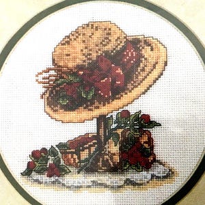 May include: A straw hat with red ribbon and berries, sitting on a wooden stand with more berries around it. The hat is decorated with green leaves and red berries.