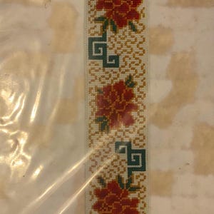 May include: A woven fabric trim featuring a repeating pattern of red floral motifs and dark green geometric shapes set against a light beige background with a textured, golden-yellow interlocking line design throughout the length of the strip.