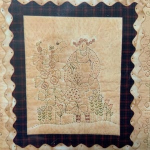 Op de afbeelding: Een beige vierkant van stof met een bruine en zwarte geruite rand. In het midden van het vierkant is een geborduurd beeld van een vrouw in een jurk die in een tuin met bloemen staat. De vrouw houdt een bloem in haar hand. De bloemen zijn geborduurd in tinten bruin, roze en groen.