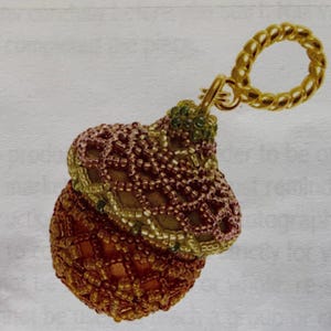 May include: A beaded acorn-shaped ornament with a gold-coloured chain loop. The ornament is primarily orange and brown, with a mesh-like pattern. The top of the acorn is green and gold. The chain loop is a twisted rope design.
