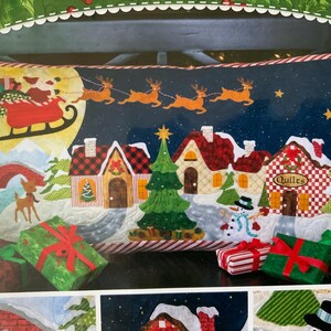 May include: A festive Christmas cushion depicting a winter village scene with Santa's sleigh, reindeer, and snow-covered houses. The cushion has a dark blue night sky background with stars and a full moon. Several wrapped gifts are in the foreground.