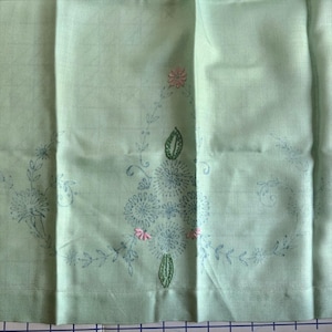 May include: A pale green fabric with embroidered floral patterns. The embroidery includes blue and pink flowers, green leaves, and fine vines. The fabric is folded, revealing the texture and design of the embroidery.