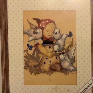 May include: Cross-stitch picture in a brown frame, depicting a child with doves. The child wears a red spotted bonnet and a yellow dress with blue dots. The artwork is framed with a cream-coloured mount.