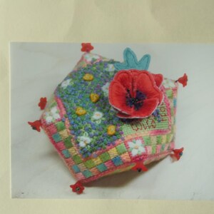 Puede incluir: Un cojín decorativo cuadrado hecho a mano con un diseño floral. El cojín presenta una amapola roja y un acento de hoja azul. El cojín tiene un diseño de patchwork con varios colores, incluyendo rosa, verde y amarillo. Acentos rojos en cada esquina.