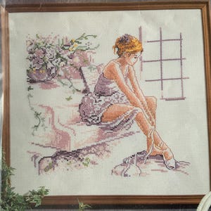 Op de afbeelding: Kruissteekkunstwerk van een ballerina die zit en haar spitsschoenen vastmaakt. Het stuk is in hout ingelijst en heeft een zacht kleurenpalet van roze, paars en crèmes. Op de achtergrond is een raam met een rasterpatroon te zien en er zijn bloemelementen aanwezig.