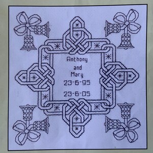 May include: A cross-stitch design featuring a central square with the names "Anthony and Mary" and the dates "23-6-95" and "23-6-05". The design is framed by an intricate knot pattern and bell-shaped ornaments with bows at each corner.
