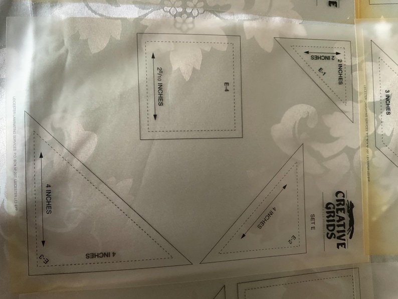 以下が含まれることがあります： 「Creative Grids Set E」とラベル付けされた透明なプラスチック製のキルティングテンプレートシート。2インチ、3インチ、4インチなどのインチ単位の測定値が印刷された三角形や正方形などのさまざまな幾何学模様が、白い背景の上に配置されています。