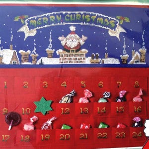 May include: A red fabric advent calendar featuring a cross-stitched blue top panel with a Santa Claus figure on a chimney and the text Merry Christmas. The calendar has twenty-four numbered pockets, some containing various wrapped candies and a lollipop.