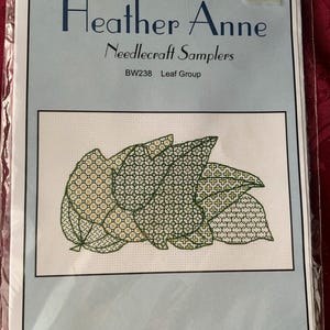 May include: A Heather Anne Needlecraft Samplers kit, BW238 Leaf Group, featuring a cross-stitch pattern of overlapping leaves with green and yellow geometric details. The package includes fabric, threads, a needle, and instructions. Design size: 16cm by 9cm.