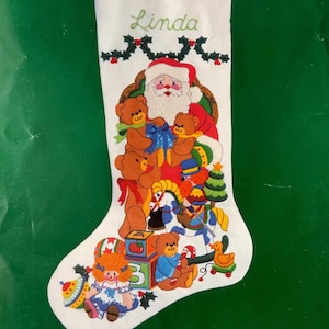May include: White Christmas stocking embroidered with Santa Claus, teddy bears, and toys. The name "Linda" is embroidered in green script at the top. The stocking is decorated with holly and berries and has a green background.