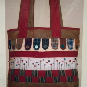 Puede incluir: Un bolso de tela rectangular con una sección superior e inferior marrón, una banda horizontal roja y un panel central blanco decorado con tallos bordados rematados con pequeños botones de colores. El bolso tiene asas rojas y un patrón.