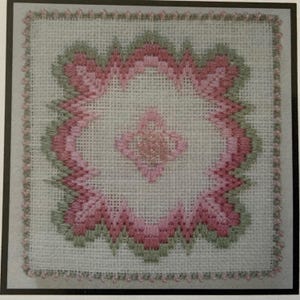 Könnte beinhalten: Ein quadratisches Stickbild auf weißem Leinen mit einem zentralen, sternförmigen rosa Motiv, das von einer größeren, komplexen geometrischen Bordüre in Rosa und Salbeigrün umgeben ist. Die Ränder sind mit kleinen rosa Perlen verziert.