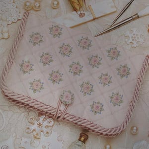 Peut inclure: Une pochette matelassée rose clair avec un motif de roses fleuries. La pochette est ornée de roses roses et d'accents bleus, et possède une bordure en corde rose. La fermeture comporte un bouton assorti en forme de rose.