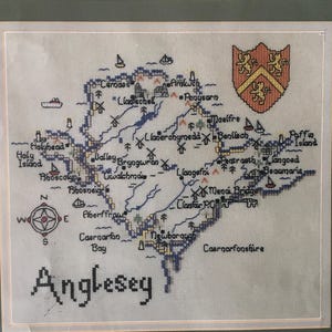 May include: A cross-stitch map of Anglesey, an island off the coast of Wales. The map is framed and features a compass, a coat of arms, and the names of various towns and villages on the island.