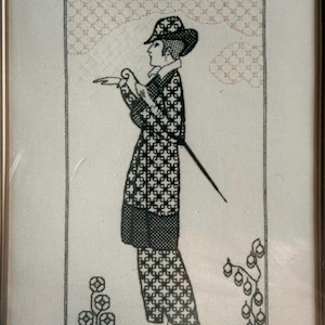 Op de afbeelding: Ingelijste zwart-wit kruissteekkunst van een persoon in een patroon outfit en hoed, met een wandelstok. De kunst bevat bloemen en geometrische ontwerpen, met een neutrale achtergrond en een goudkleurige lijst.