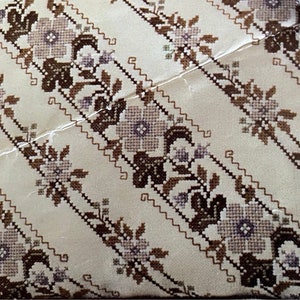 Könnte beinhalten: Ein weißes Stoffkissen mit einem braunen und violetten Stickmuster mit Blumen. Das Kissen ist mit einem sich wiederholenden Muster aus Blumen und Blättern verziert.