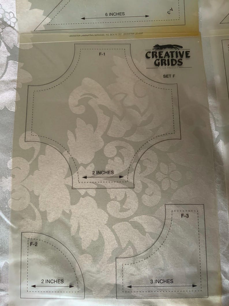 以下が含まれることがあります： Creative Grids Set Fとラベル付けされた透明なプラスチック製のキルティングテンプレートシート。シートにはF-1、F-2、F-3とマークされた3つの異なる形状があり、2インチと3インチの測定値が印刷されています。テンプレートには黒い輪郭とテキストが含まれています。