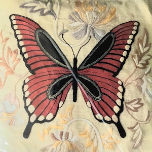 May include: Embroidered butterfly design with red wings, black accents, and white spots. The butterfly is centered on a cream-colored background with floral embroidery in various colors. The image showcases a detailed textile art piece.