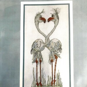 May include: Cross stitch pattern featuring two grey flamingos with their necks intertwined to form a heart shape. The flamingos are standing on long, thin legs in a grassy area.