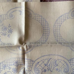 May include: A beige fabric with blue embroidery designs. The designs are of a grid pattern with floral accents. The fabric is folded in half.