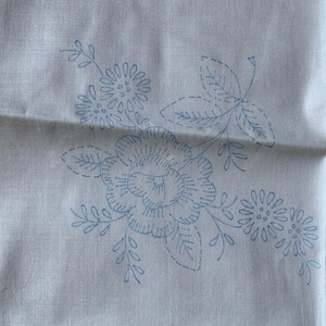 Könnte beinhalten: Ein weißer Leinenstoff mit blauen gestickten Blumen. Das Design zeigt eine große Rose mit kleineren Blumen und Blättern.