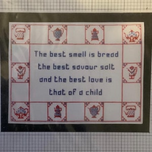 Könnte beinhalten: Ein gerahmtes Kreuzstichstück mit dem Text "The best smell is bread, the best savour salt, and the best love is that of a child." Das Design zeigt einen weißen Hintergrund mit roten und blauen Kreuzstichrändern und dekorativen Motiven.