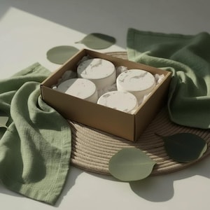 May include: A square cardboard gift box containing four round, white, marble-patterned coasters. The coasters are nestled in white tissue paper within the box, which is surrounded by sage green fabric and several loose, flat, green eucalyptus leaves.