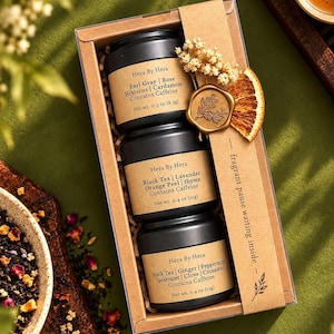May include: A gift set of three black cylindrical tea tins labelled Hera By Hera, featuring ingredients like Earl Gray, rose, hibiscus, cardamom, black tea, lavender, orange peel, thyme, ginger, peppermint, spearmint, clove, and cinnamon.