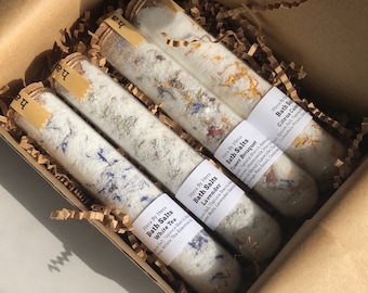 May include: Four glass tubes filled with bath salts and dried flowers. The tubes are labeled with the names of the scents: White Tea, Lavender, Flower Bouquet, and Citrus Cake. The tubes are in a brown cardboard box with shredded paper.