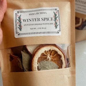 Puede incluir: Una bolsa de papel marrón que contiene rodajas de naranja secas, hojas de laurel y especias. La etiqueta dice HERA BY HERA WINTER SPICE STOVETOP SIMMER POTPOURRI Net Wt. 3 OZ (85 g) con un borde decorativo de bayas rojas y hojas verdes.