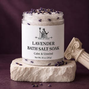 May include: A clear plastic jar filled with white bath salts and dried purple lavender buds. The white label features the text HERA BY HERA, LAVENDER BATH SALT SOAK, Calm & Unwind, and Net Wt: 10 oz (283g) with a black line drawing of a goddess.