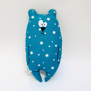 Peut inclure: Un jouet en peluche bleu en forme d'ours avec des étoiles blanches imprimées sur le tissu. L'ours a un nez et une bouche brodés noirs et de grands yeux blancs. Le jouet porte une étiquette qui dit "L'AMITOU".