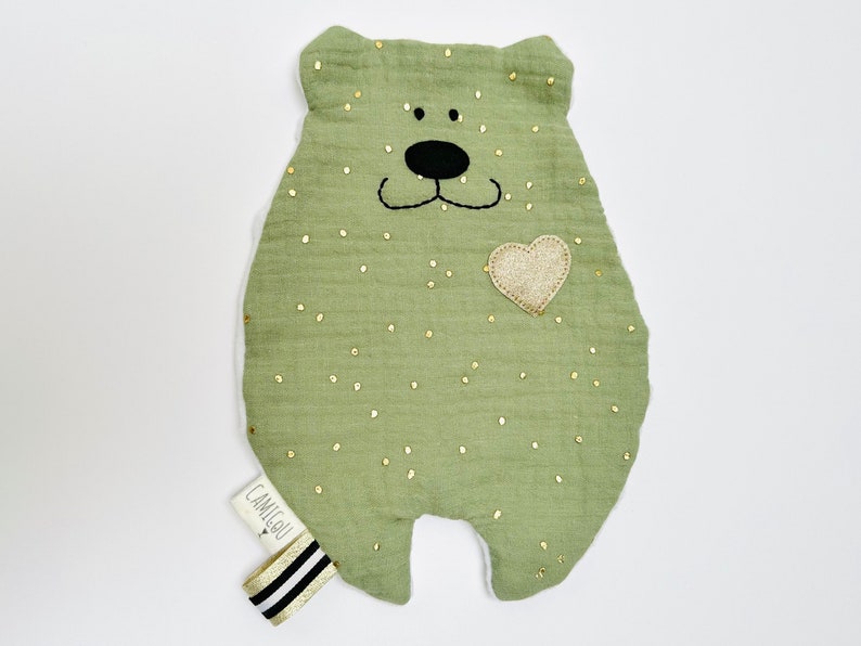 May include: A green plush bear with gold glitter dots and a gold heart on its chest. The bear has a black embroidered face and the text "CAMIGOU" is on the bottom left corner.