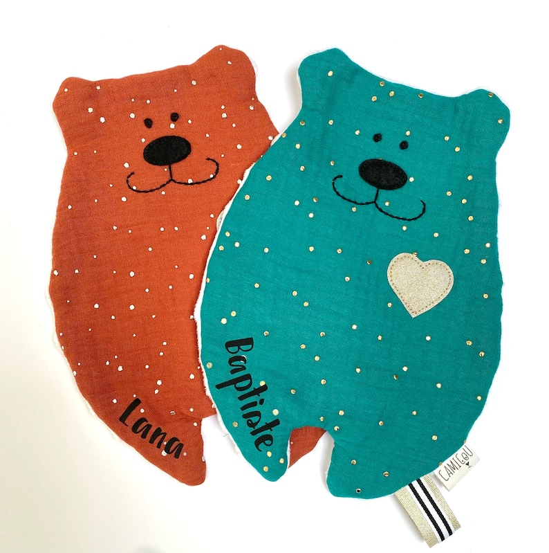 May include: Two plush bear-shaped baby blankets with embroidered names. One is teal with gold glitter dots and a gold heart, the other is rust with white glitter dots. Both have black embroidered eyes, noses, and mouths.