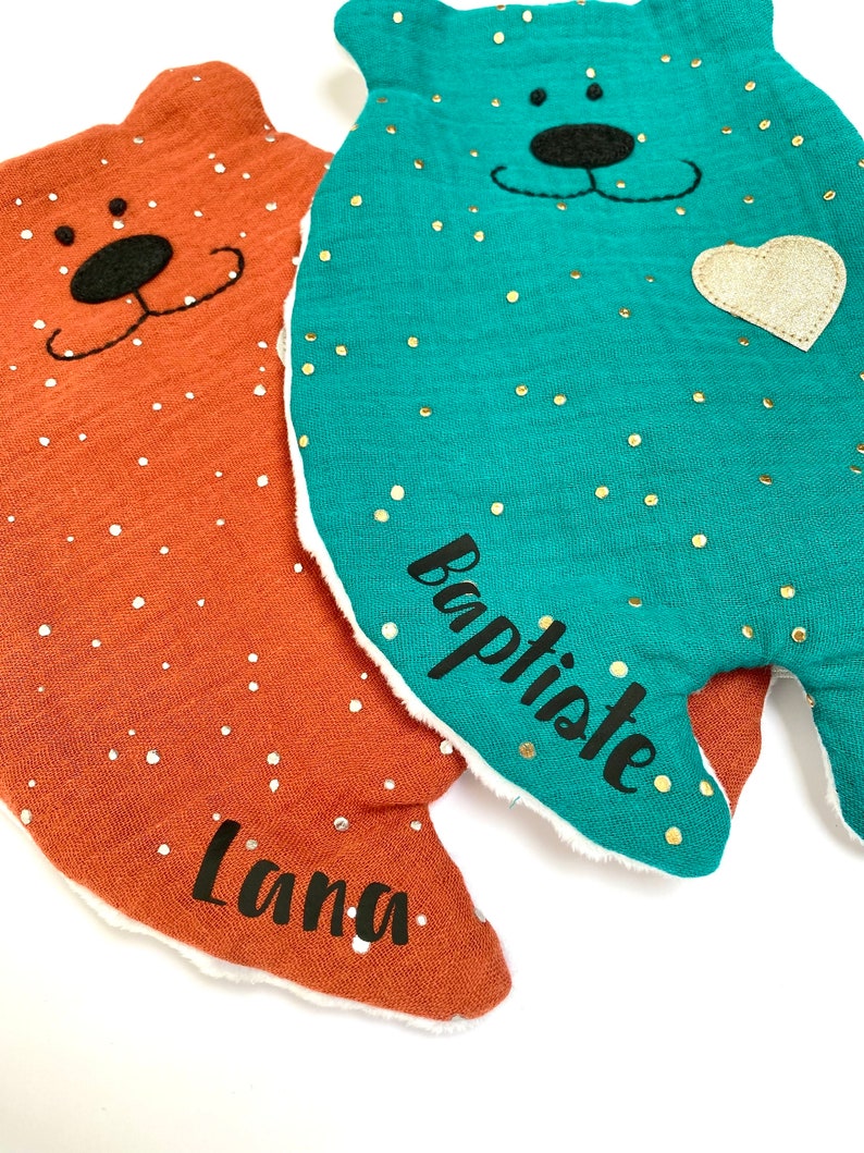 May include: Two plush bear-shaped baby blankets with embroidered faces. The blanket on the left is orange with white polka dots and the name "Lana" embroidered on it. The blanket on the right is teal with gold polka dots and the name "Baptiste" embroidered on it.