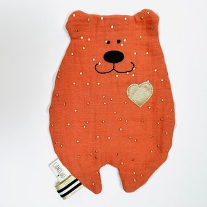Puede incluir: Un oso de peluche suave, de color naranja, con detalles de purpurina dorada y un corazón dorado en el pecho. El oso tiene una nariz y una boca bordadas en negro. El oso está etiquetado con "Camigu" en la parte inferior.