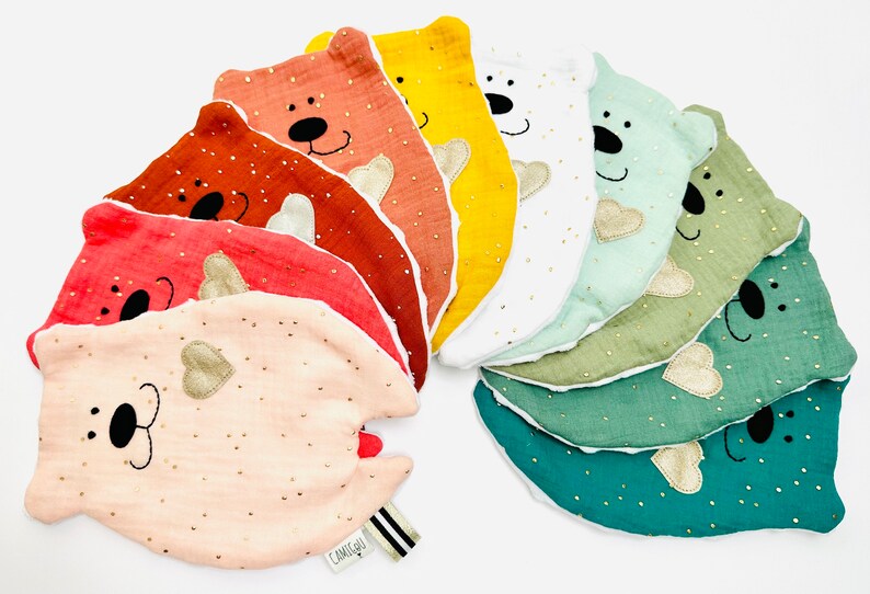 May include: A set of ten colourful, soft, plush bear-shaped baby toys with embroidered faces and gold hearts. The toys are arranged in a rainbow pattern.