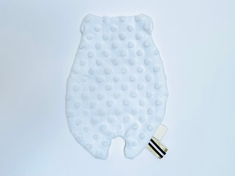 May include: White plush baby toy shaped like a bear with a textured surface. The toy has a small white tag with black and gold stripes attached to it.
