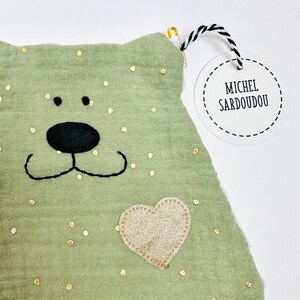 May include: A green fabric pouch with gold glitter dots, a black embroidered bear face, and a gold glitter heart. The pouch has a white tag attached with black text that reads "Michel Sardoudou".