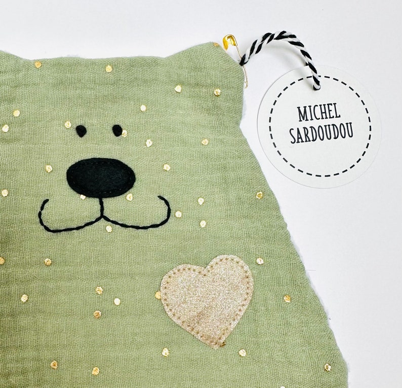 May include: A green fabric pouch with gold glitter dots, a black embroidered bear face, and a gold glitter heart. The pouch has a white tag attached with black text that reads "Michel Sardoudou".