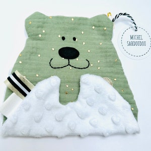 May include: A green plush bear with a white, textured tummy and gold glitter accents. The bear has black embroidered eyes and a nose. A white tag with black text reads "Michel Sardou"