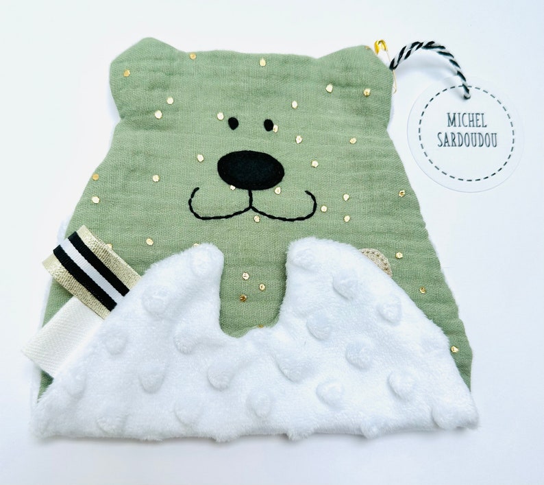 May include: A green plush bear with a white, textured tummy and gold glitter accents. The bear has black embroidered eyes and a nose. A white tag with black text reads "Michel Sardou"