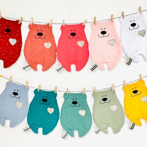 May include: A string of ten plush bear toys, each with a different colour and pattern. The bears are hanging from a clothesline with clothes pegs. Each bear has a small heart on its chest.