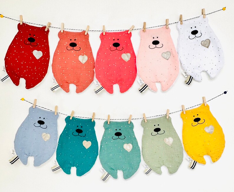 May include: A string of ten plush bear toys, each with a different colour and pattern. The bears are hanging from a clothesline with clothes pegs. Each bear has a small heart on its chest.