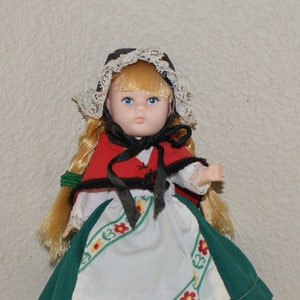 May include: A porcelain doll wearing a green and white dress with a red jacket and a white lace bonnet. The doll has blonde hair and blue eyes.