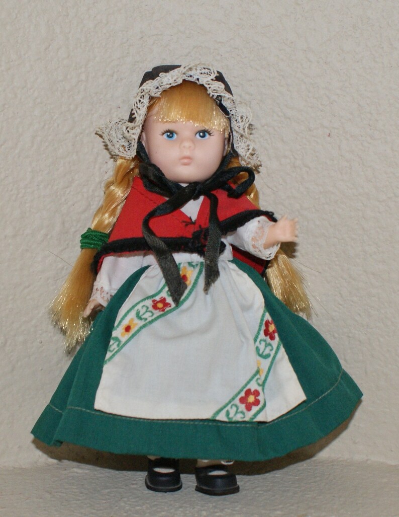 May include: A porcelain doll wearing a green and white dress with a red jacket and a white lace bonnet. The doll has blonde hair and blue eyes.