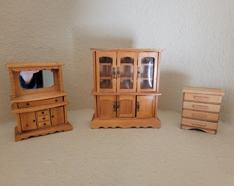 May include: Three miniature wooden cabinets, one with glass doors and shelves, one with a mirror and drawers, and one with drawers. The cabinets are all made of light wood and have a rustic finish.